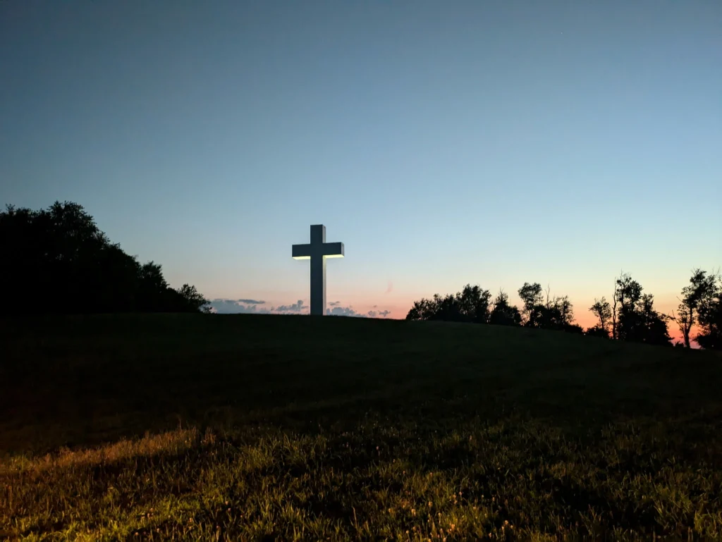 White cross on a grassy hill at dusk, with a line of trees along the horizon and a gradient sky from orange to deep blue.