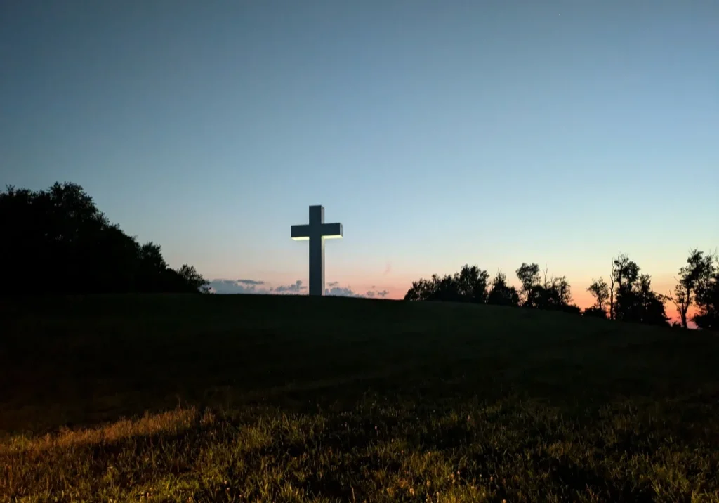 White cross on a grassy hill at dusk, with a line of trees along the horizon and a gradient sky from orange to deep blue.